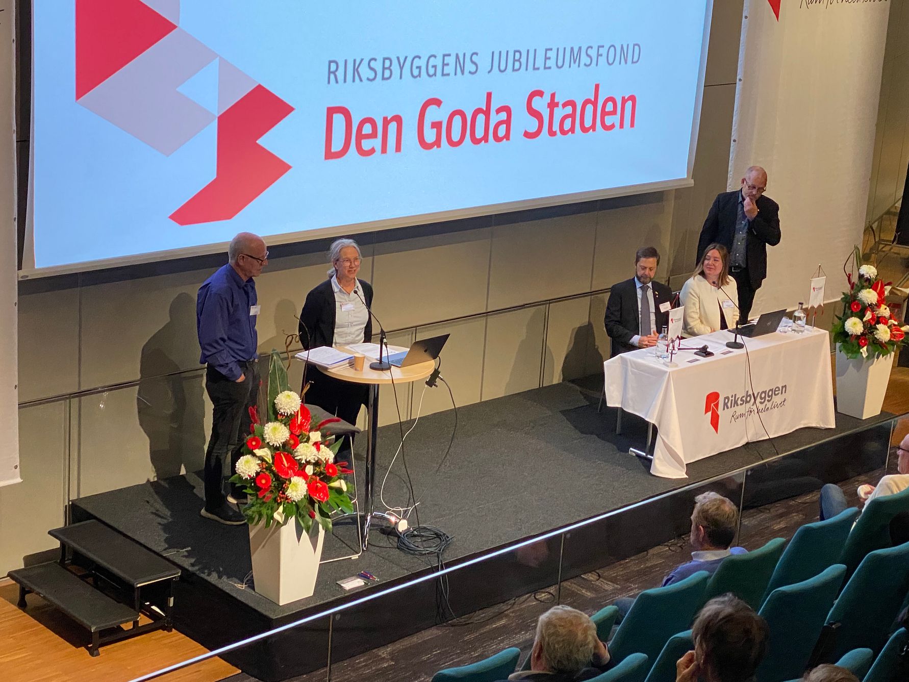 Föreläsning i en konferenslokal med en panel på scenen, blommor vid podierna och en stor skärm som visar texten "Riksbyggens jubileumsfond - Den Goda Staden