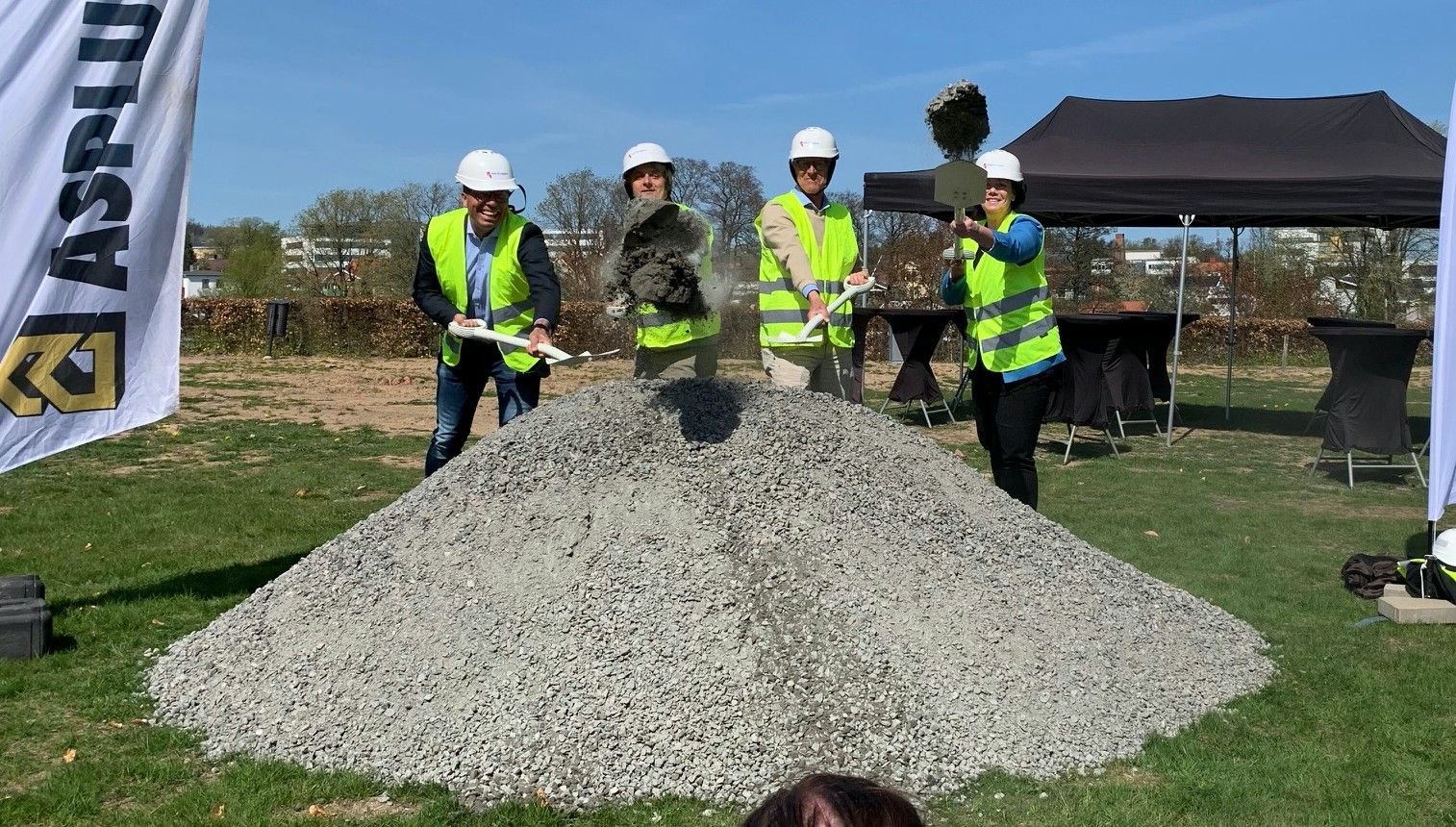 Fyra personer i reflexvästar och hjälmar använder spadar för att symboliskt vända grus vid en utomhusceremoni, med en hög grus i förgrunden och tält i bakgrunden