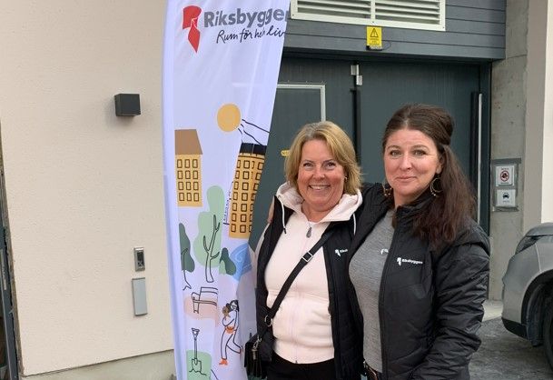 Två personer står framför en byggnad bredvid en flagga med Riksbyggens logotyp och texten "Rum för hela livet
