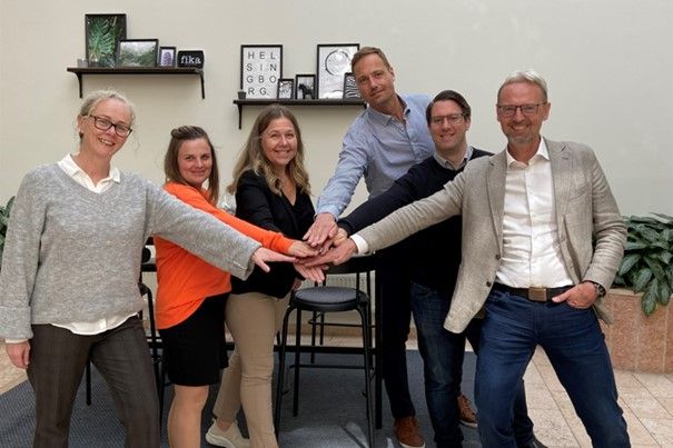 En grupp människor står i en cirkel och lägger sina händer tillsammans i mitten, i en inomhusmiljö med skrivbord, stolar och växter i bakgrunden