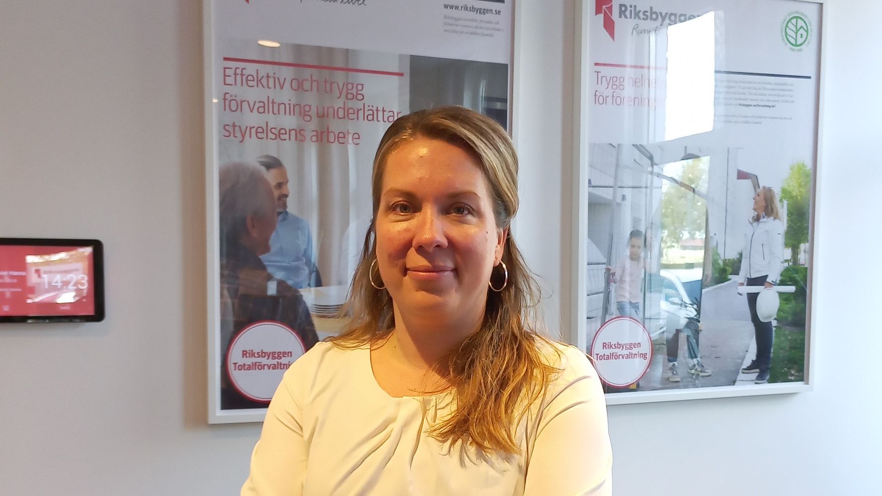 Kvinna i vit topp står inomhus framför två affischer och ett fönster med utsikt mot en grönskande omgivning