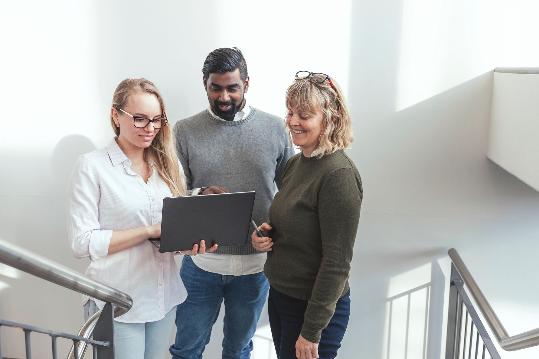 Tre personer står på en trappa och tittar på en laptop, med ljusinsläpp från fönster i bakgrunden