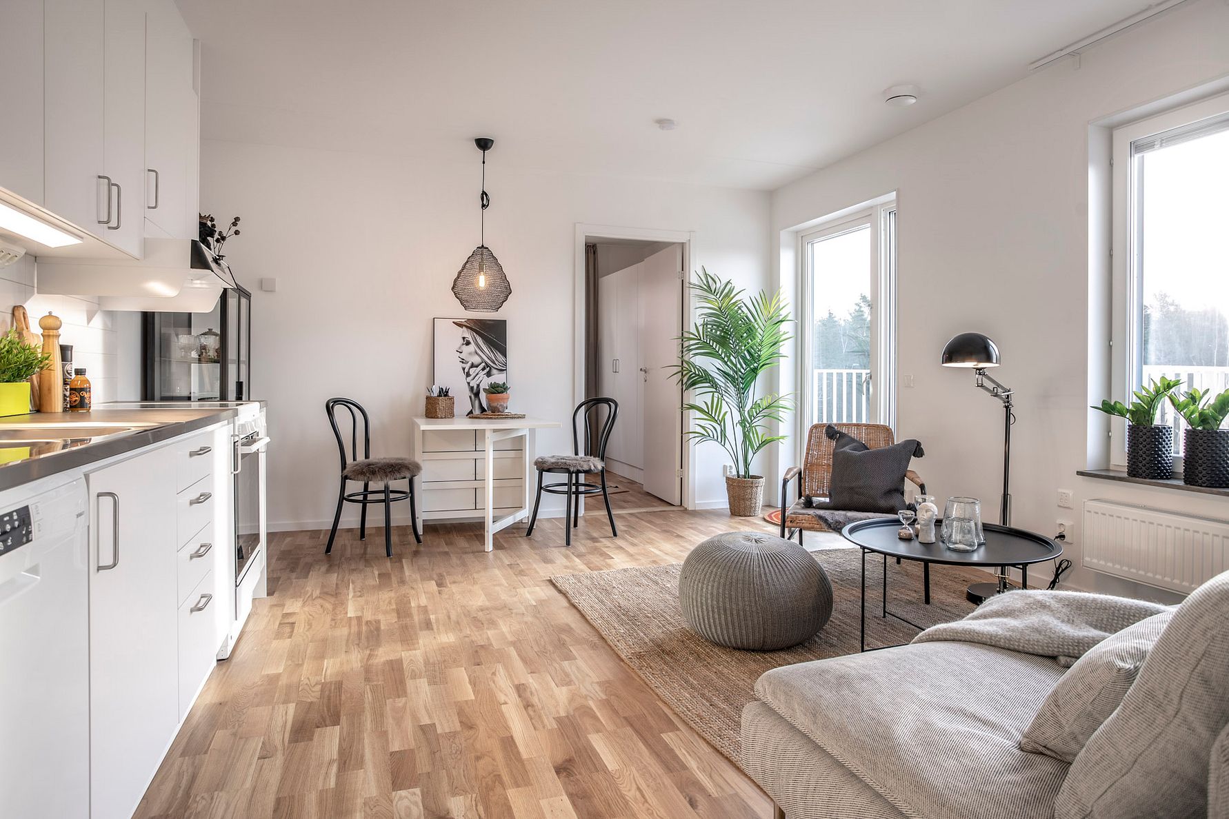 Ljust vardagsrum med parkettgolv, en grå soffa, rund sittpuff och matbord med två stolar. Dekorativ belysning, gröna växter och fönster som släpper in naturligt ljus