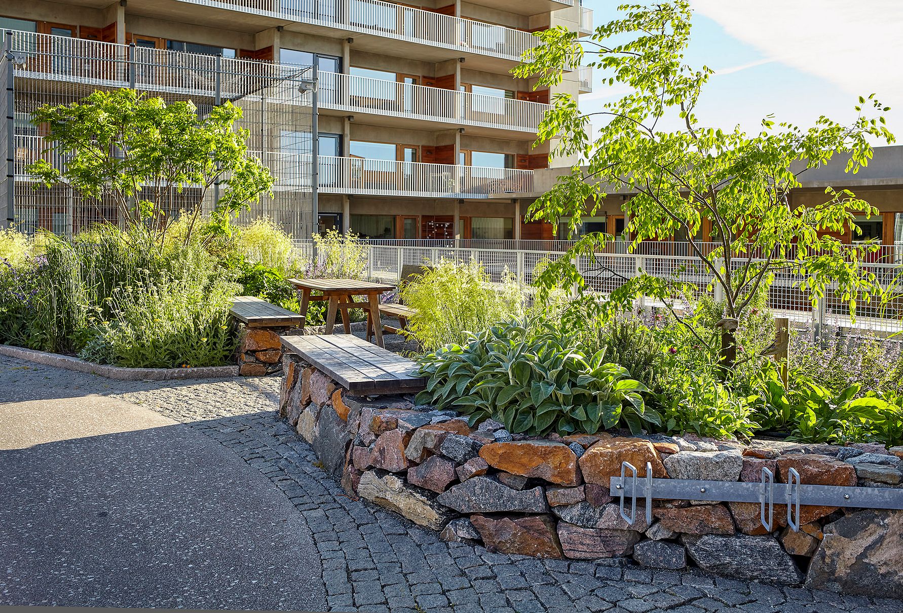 Lägenhetsbyggnad med en grönskande innergård som innehåller stenrabatter, träd, buskar och en träbänk