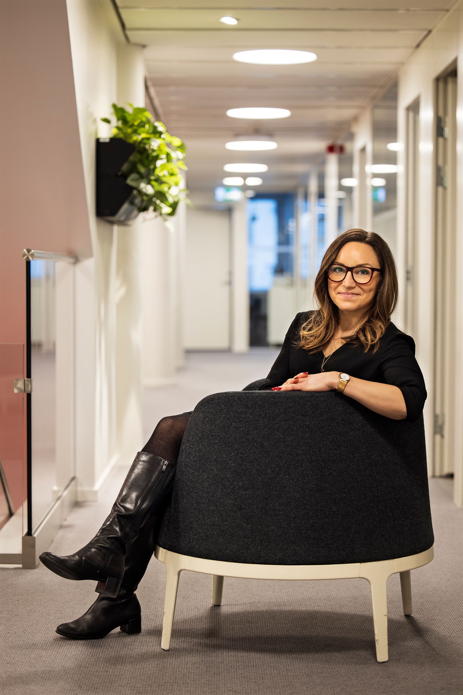 En person sitter i en mörkgrå stol i en ljus kontorskorridor med glasdetaljer och växter på väggen