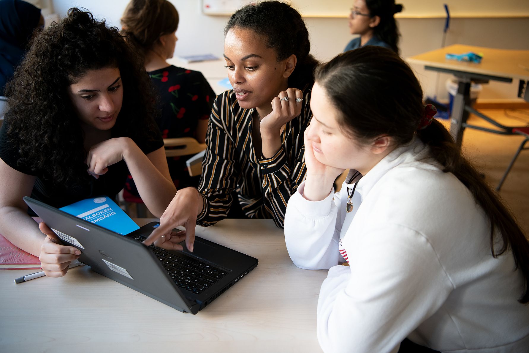Tre personer sitter runt ett bord och diskuterar medan de tittar på en bärbar dator i ett klassrum
