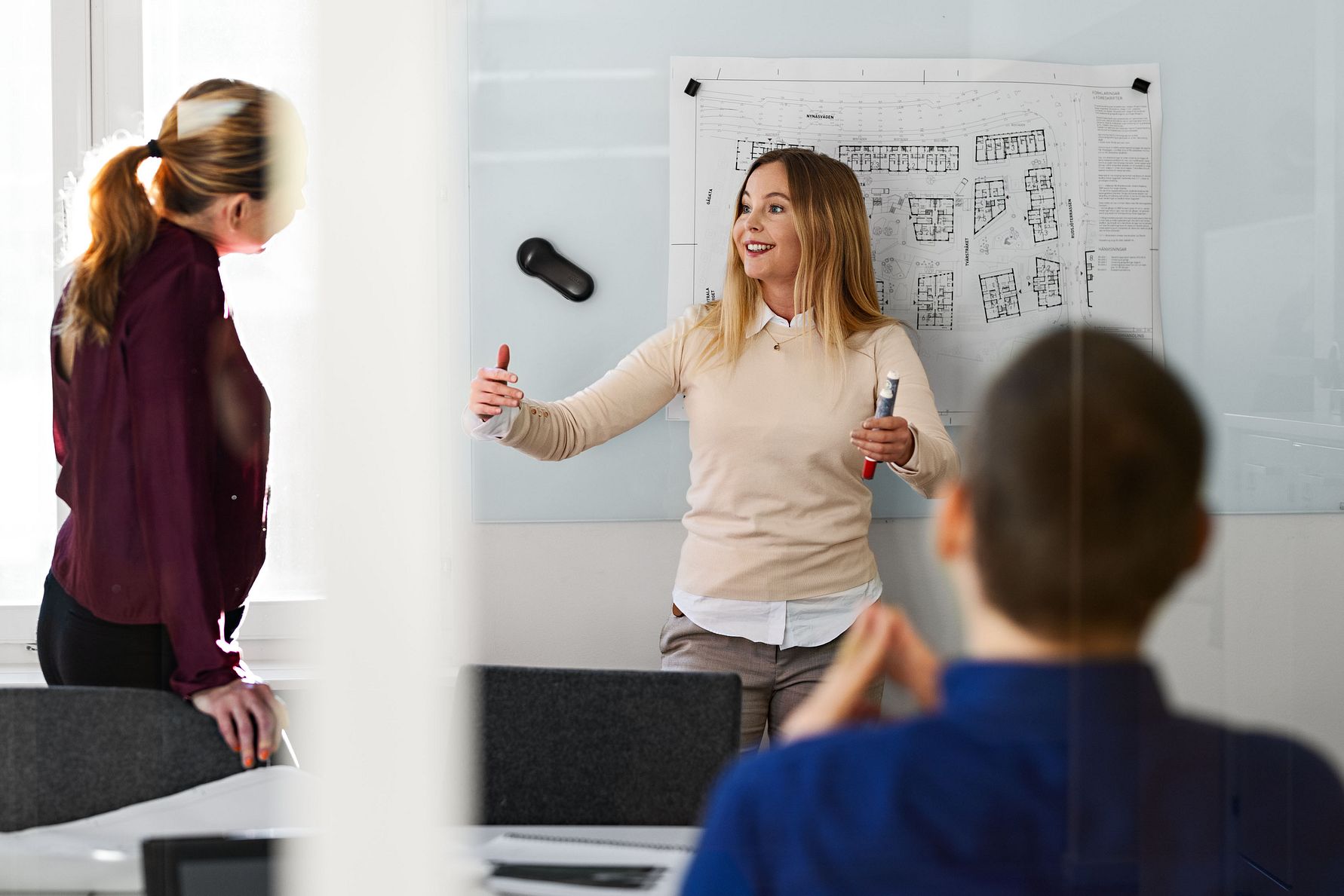 Två personer står i ett mötesrum, en kvinna håller en presentation framför en whiteboard med ritningar, medan en åhörare sitter med ryggen mot kameran