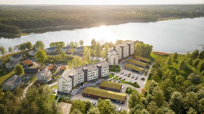 Flygvy över ett bostadsområde med moderna byggnader, parkeringsplatser och gröna tak, omgivet av skog och en sjö i bakgrunden