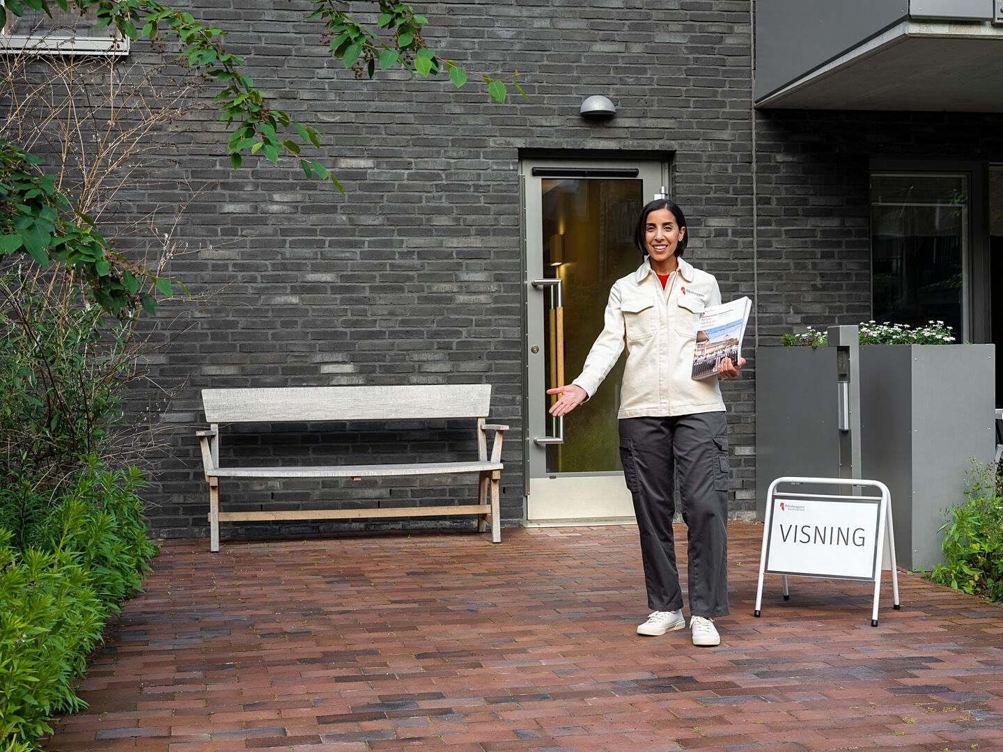 En person i vit jacka står på en stenlagd gång framför en byggnadsentré med en skylt som indikerar visning. En träbänk och växtlighet syns i bakgrunden