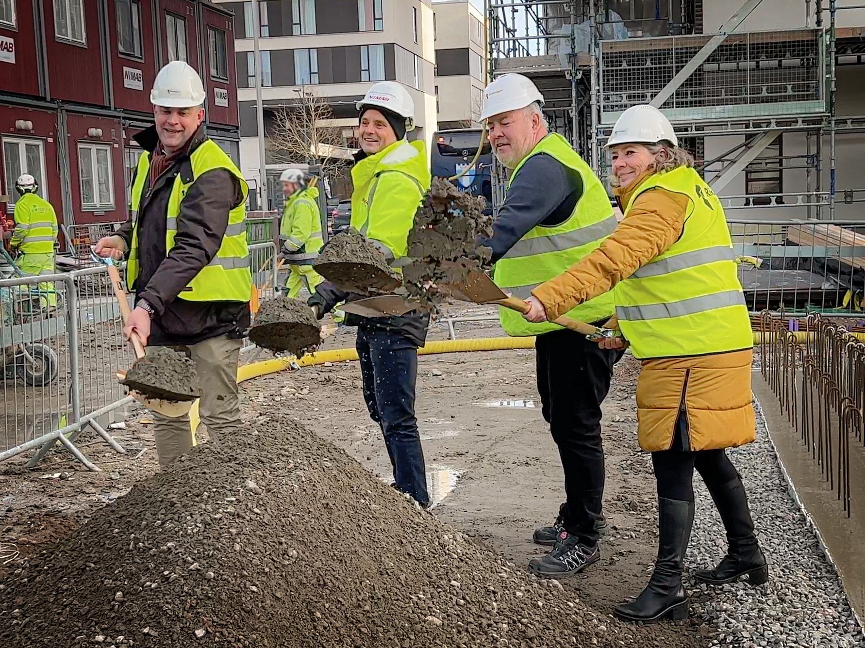 Personer i reflexvästar och bygghjälmar skyfflar jord vid en byggarbetsplats