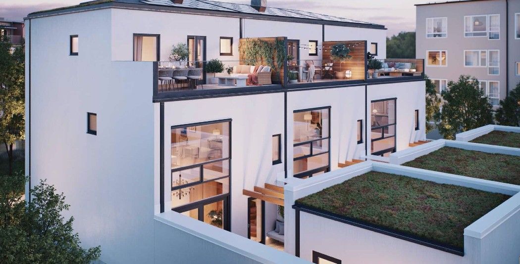 Modern vit byggnad med stora fönster och takterrass, omgiven av grönska och andra bostadshus