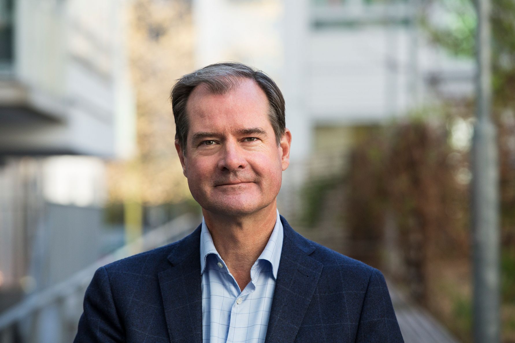 Man i blå kavaj och ljusblå skjorta står utomhus framför en suddig bakgrund av byggnader och träd