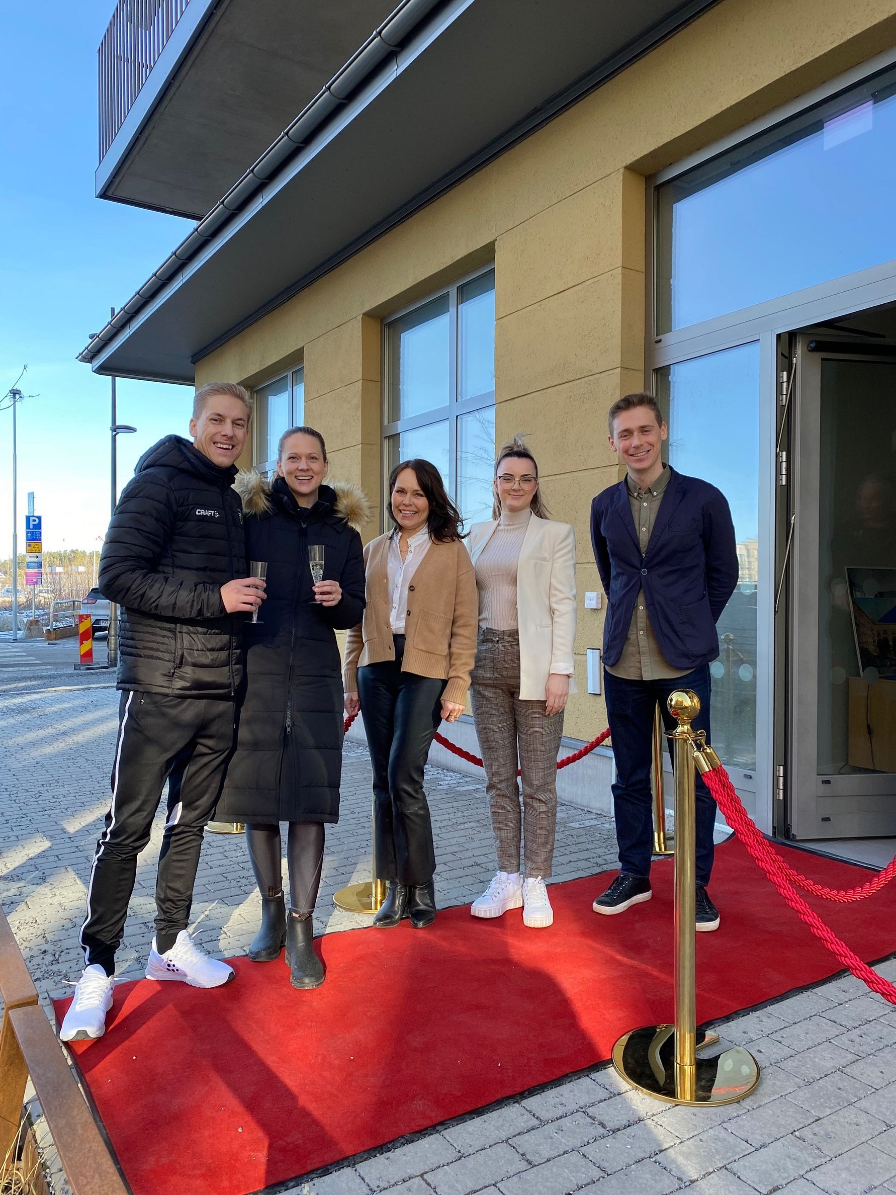 En grupp människor står på en röd matta utomhus framför en byggnad med glasdörrar, klädda i olika vinterkläder och skor