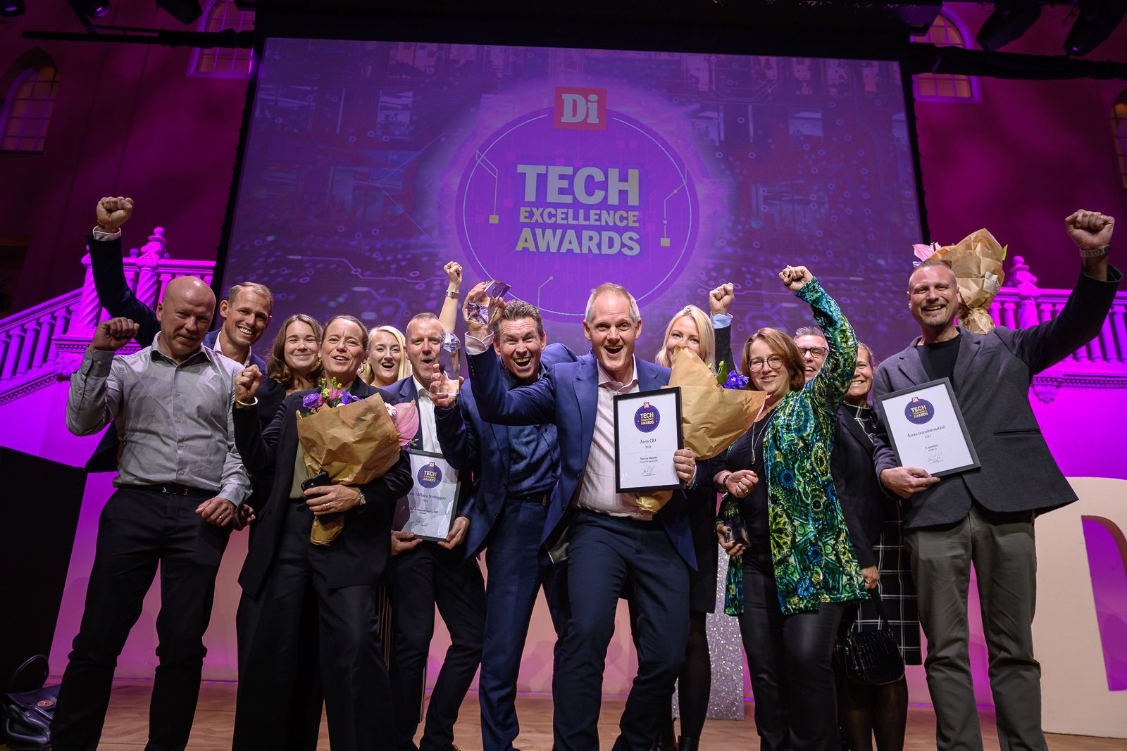 Gruppfoto av personer som firar på scen med diplom och blombuketter under Tech Excellence Awards, framför en belyst bakgrund med prislogotypen