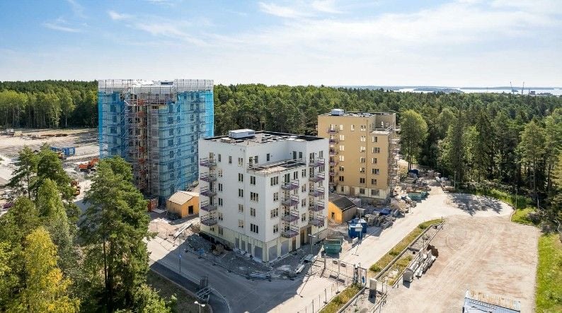 Byggarbetsplats med flerbostadshus under konstruktion, omgiven av skog och öppen mark, med klarblå himmel i bakgrunden