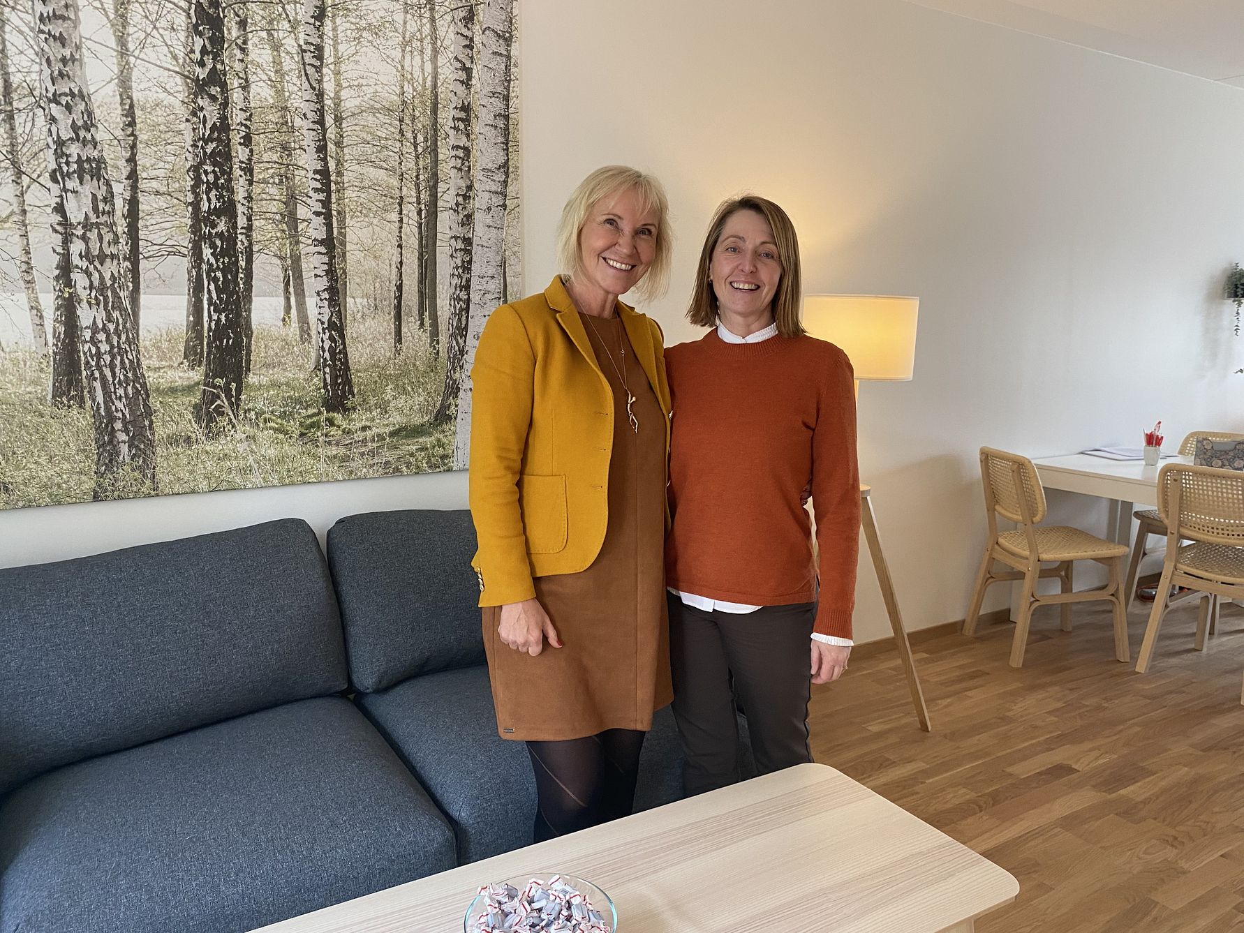 Två personer står i ett vardagsrum bredvid en mörkgrå soffa och ett soffbord med en skål godis. I bakgrunden syns en stor tavla med ett skogsmotiv, en golvlampa och matbord med stolar