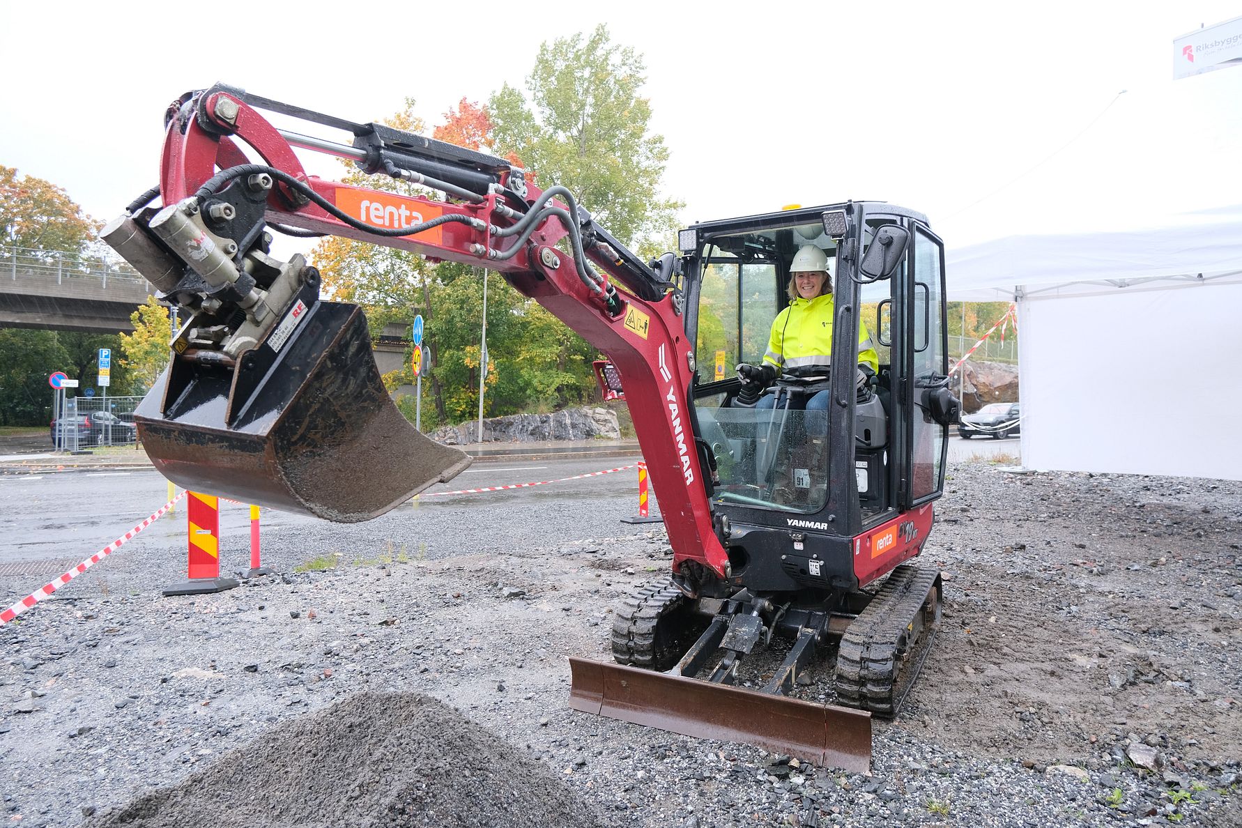 En grävmaskin på en grusplan i arbete, med en person i förarhytten. Bakgrunden visar träd och en avstängd väg med varningsmarkeringar