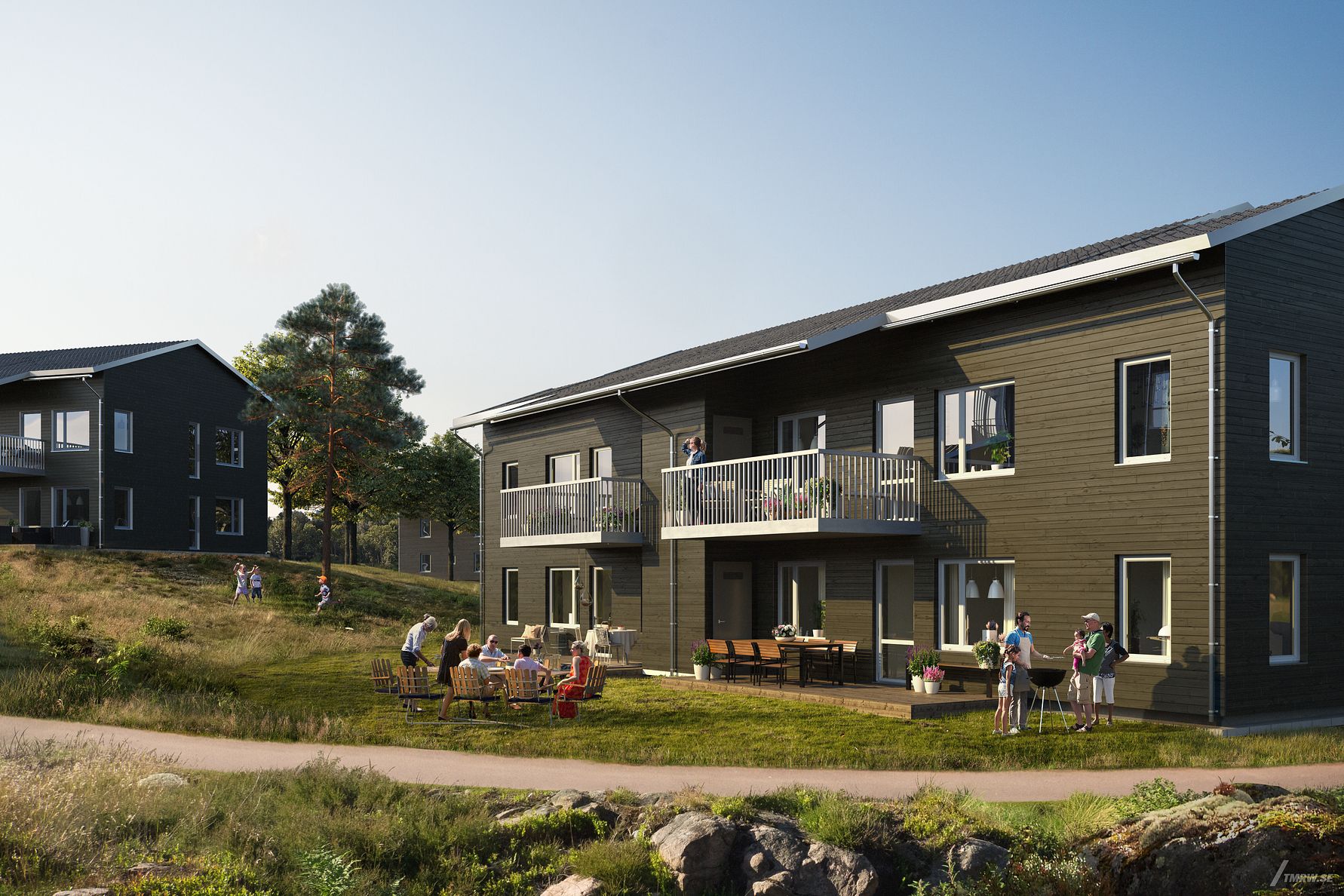 Två moderna bostadshus med mörk träfasad på en grönskande plats, personer samlade vid ett bord och barn som leker utomhus i dagsljus