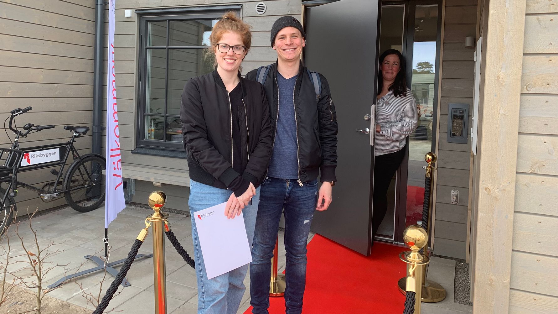 Två personer står på en röd matta utanför en byggnad med en öppen dörr. En flagga med texten "Välkommen" och en cykel syns i bakgrunden