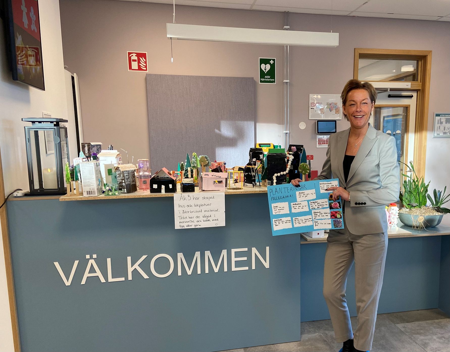 Reception med en blå disk där ordet "VÄLKOMMEN" står skrivet, omgiven av informationslappar, dekorationer och planteringar. En person i ljus klädsel står bredvid och håller i en skylt