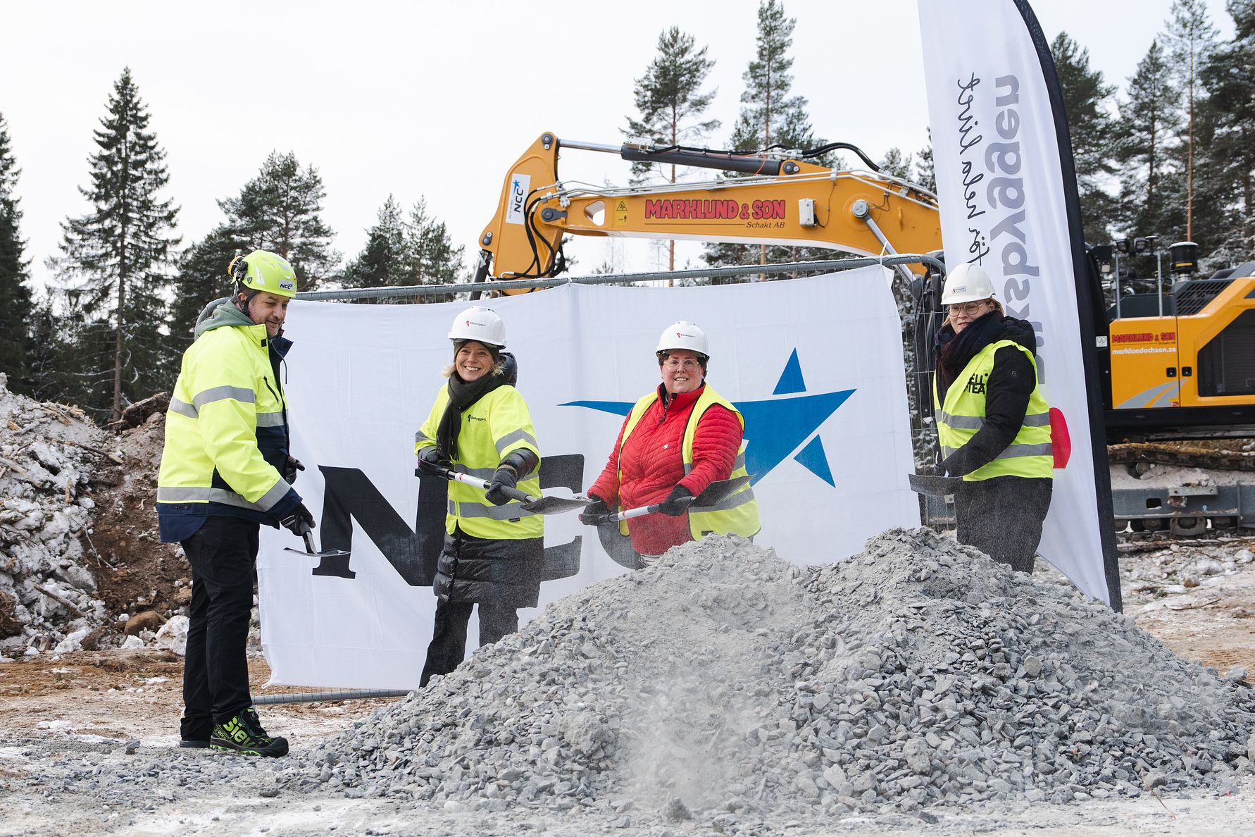 Personer med skyddsutrustning vid en byggarbetsplats, framför en grushög och en grävmaskin, med en företagsbanner i bakgrunden