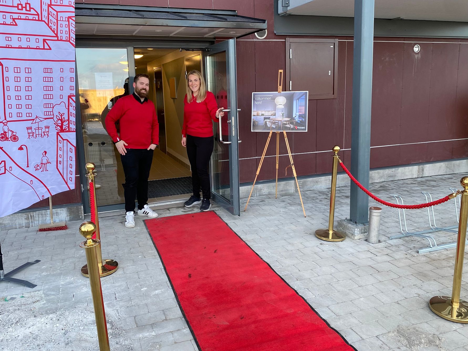 Två personer i röda tröjor står vid en ingång med en röd matta och avspärrningar. Bredvid dem står ett staffli med en affisch och i bakgrunden syns en byggnad