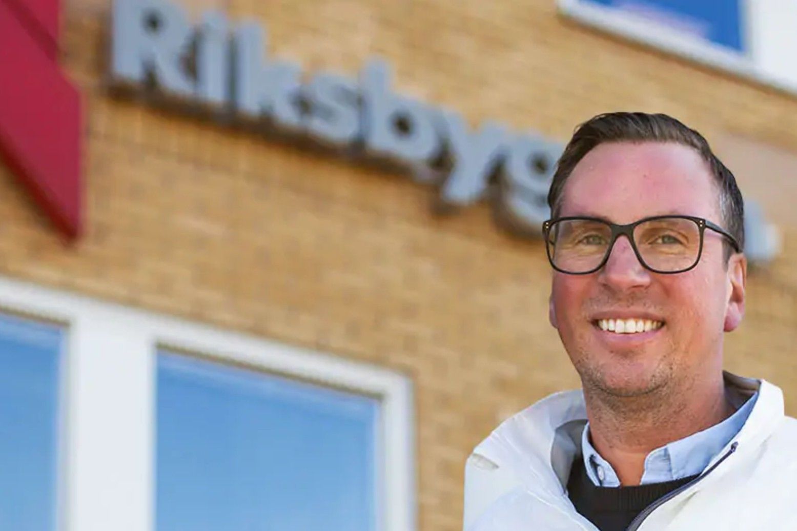 Man i vit jacka framför en byggnad med en skylt där det står "Riksbyggen