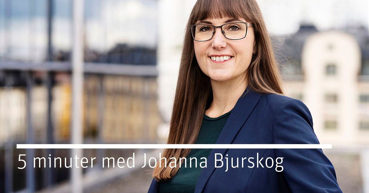 En person i en mörkblå kavaj och grönt plagg står utomhus, med en modern byggnad och stadsmiljö i bakgrunden. Texten "5 minuter med Johanna Bjurskog" syns i bild