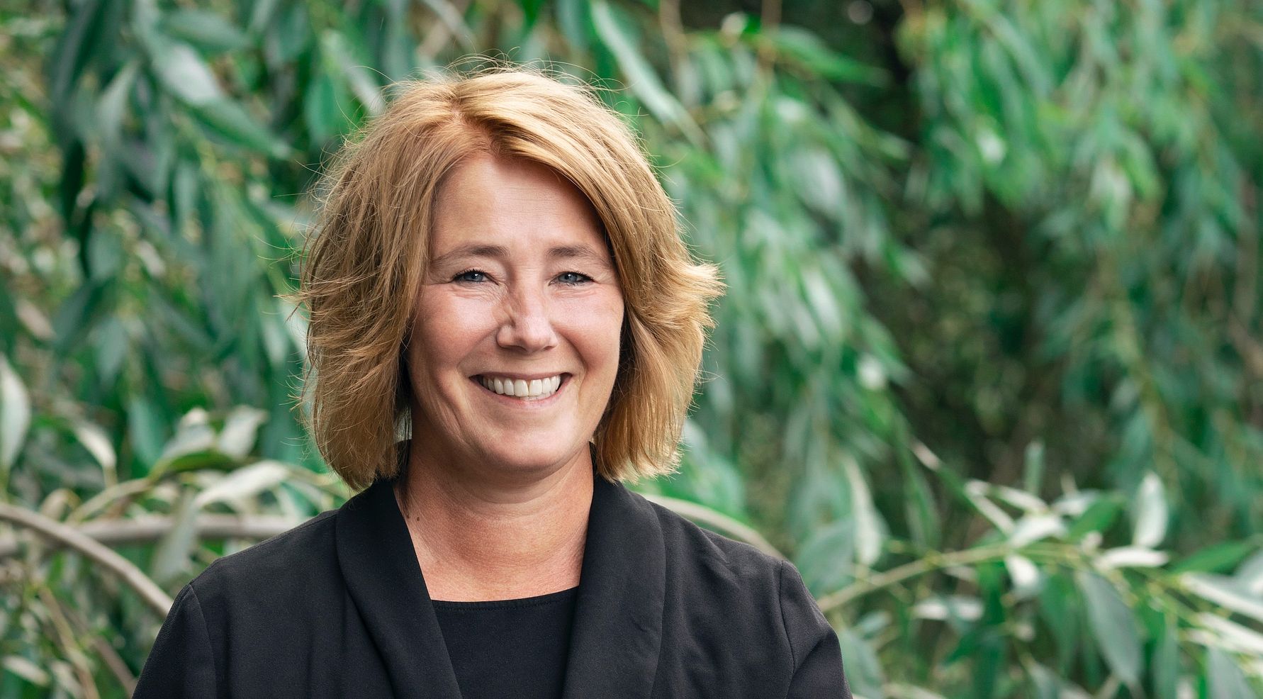 Kvinna med axellångt blont hår iförd en svart kavaj, stående framför en bakgrund av grönskande träd och bladverk