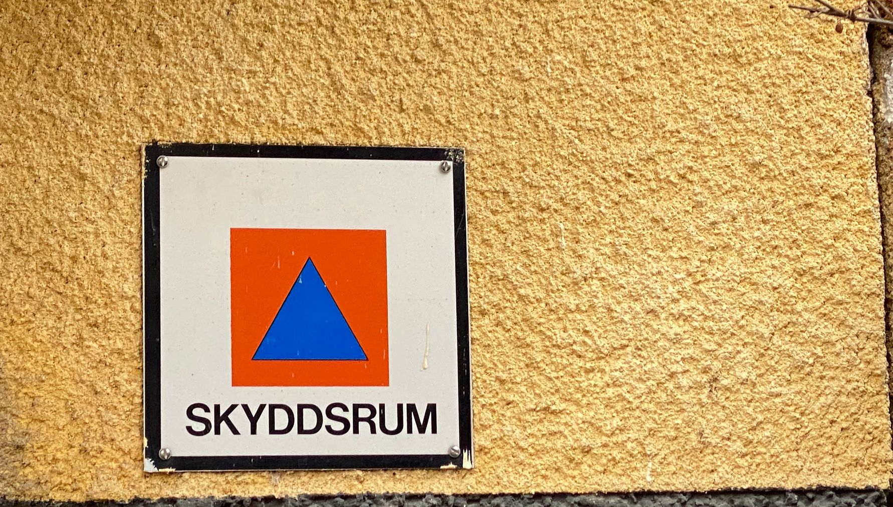 Skylt med texten "Skyddsrum" och en orange fyrkant med en blå triangel, monterad på en gul vägg