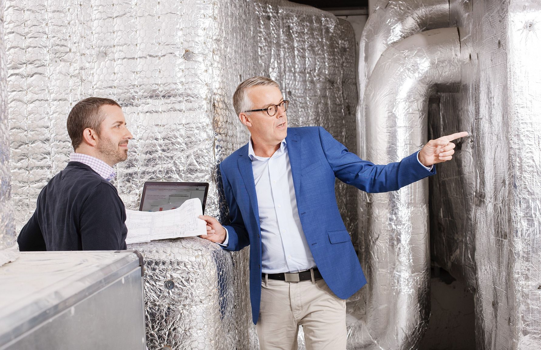 Två personer står i ett rum med isolerade rör och ventilationssystem, medan en av dem pekar och håller i ett papper