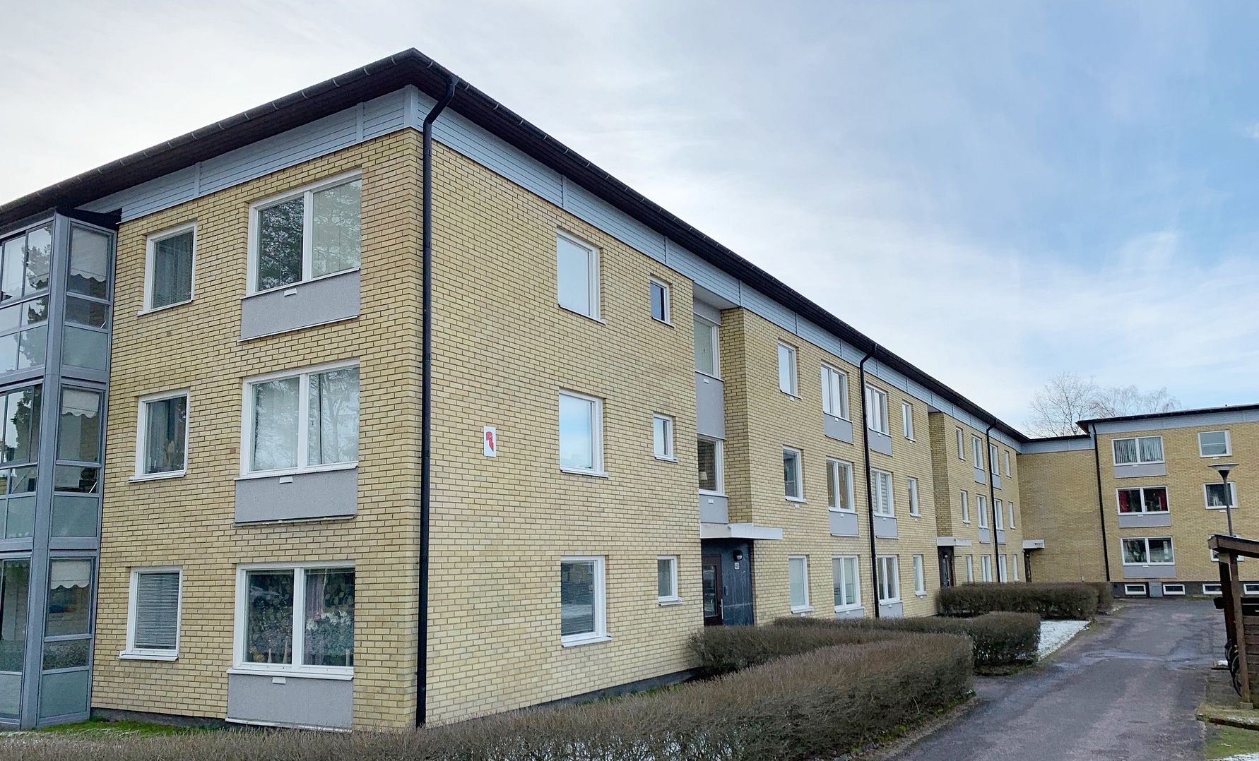 Gult flerfamiljshus med tre våningar, omgiven av en häck och en asfalterad gång