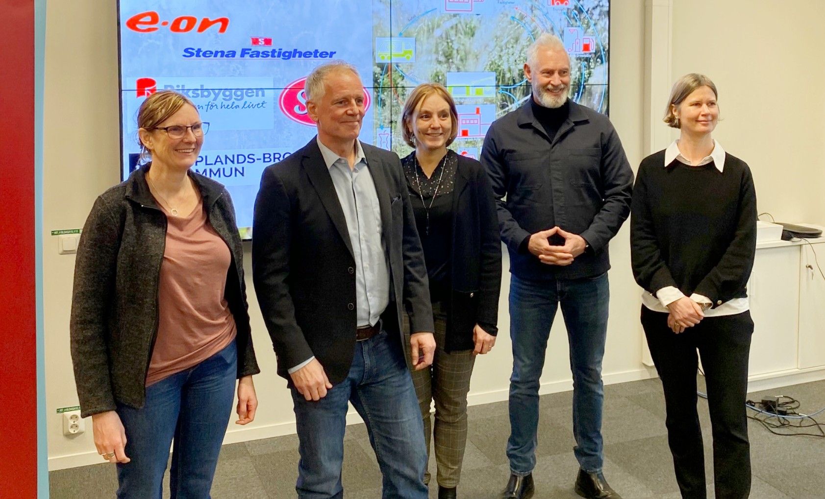 Fem personer står framför en digital skärm med företagslogotyper och text, inomhus i en kontorsmiljö. Två personer bär kavajer och andra är klädda i vardagliga kläder