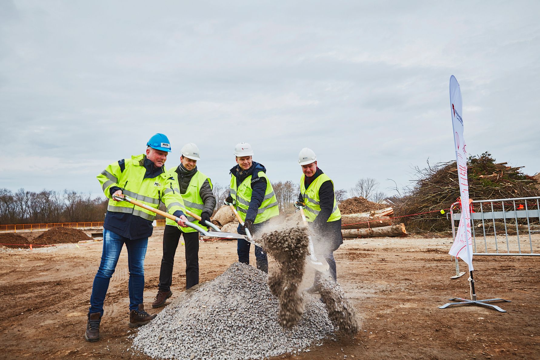 Fyra personer i varselkläder och hjälmar skyfflar grus vid en byggarbetsplats under en mulen himmel, med en reklamflagga och avverkade träd i bakgrunden