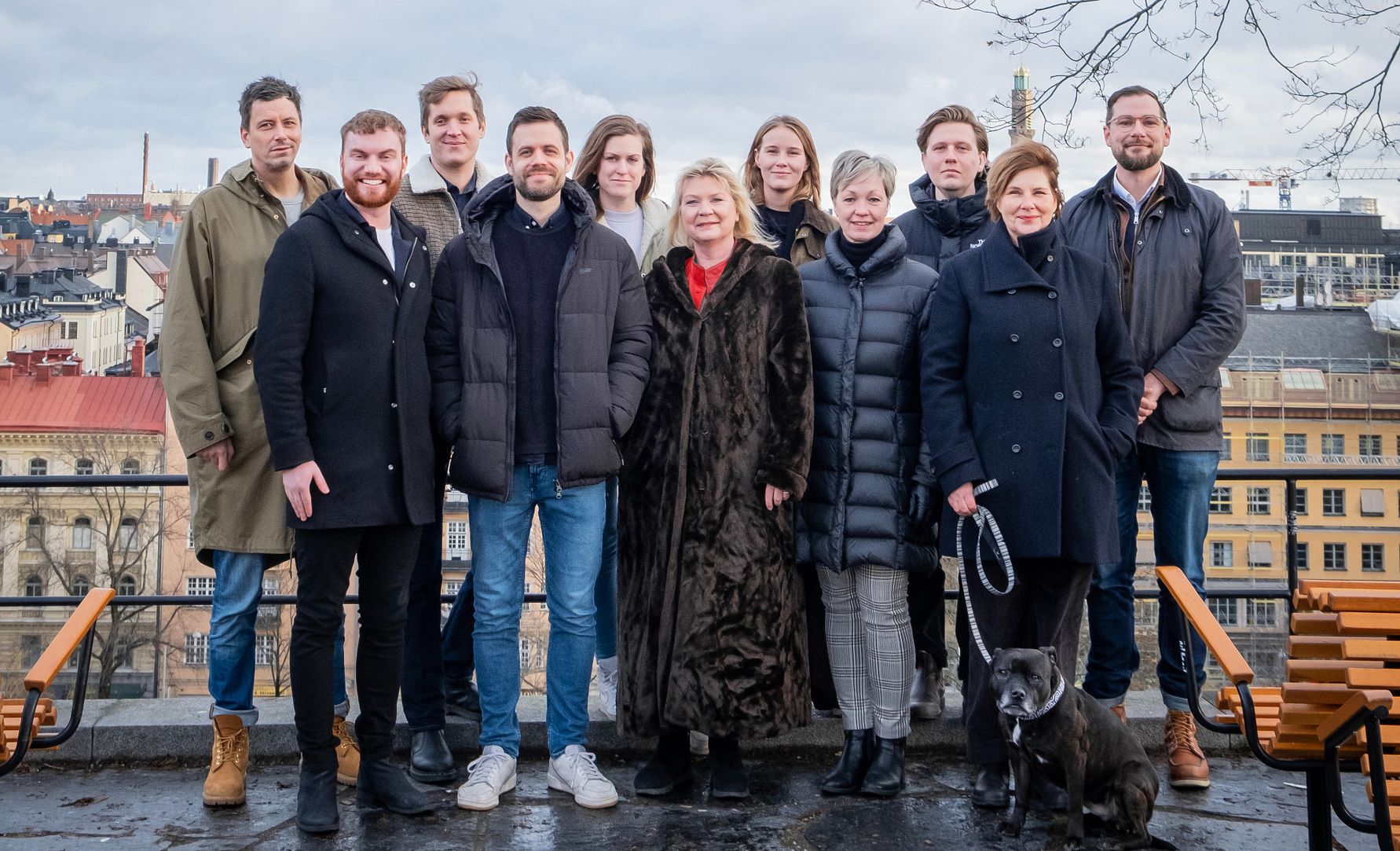 En grupp människor klädda i vinterkläder står utomhus framför en stadsutsikt med byggnader och träd i bakgrunden. En hund med koppel syns vid sidan av gruppen