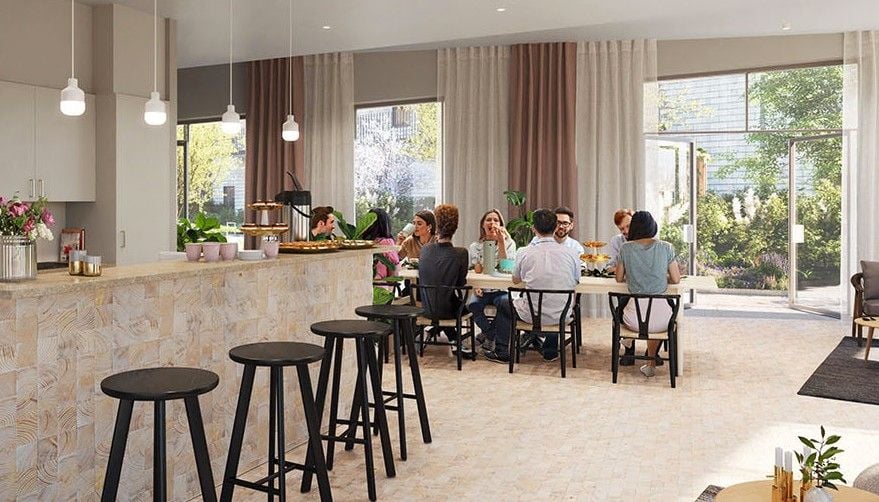 Ett modernt kök och matplats med en lång matbord där flera personer sitter och äter. Ljusa färger, barstolar vid en köksö och stora fönster som släpper in naturligt ljus