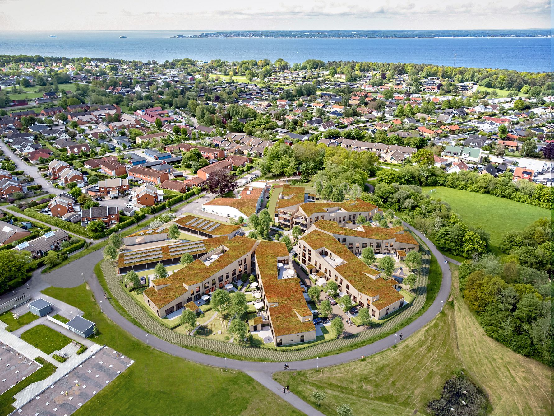 Flygbild av ett modernt bostadsområde med grön takbeklädnad, omgiven av grönska och villabebyggelse, nära en kustlinje med hav i bakgrunden