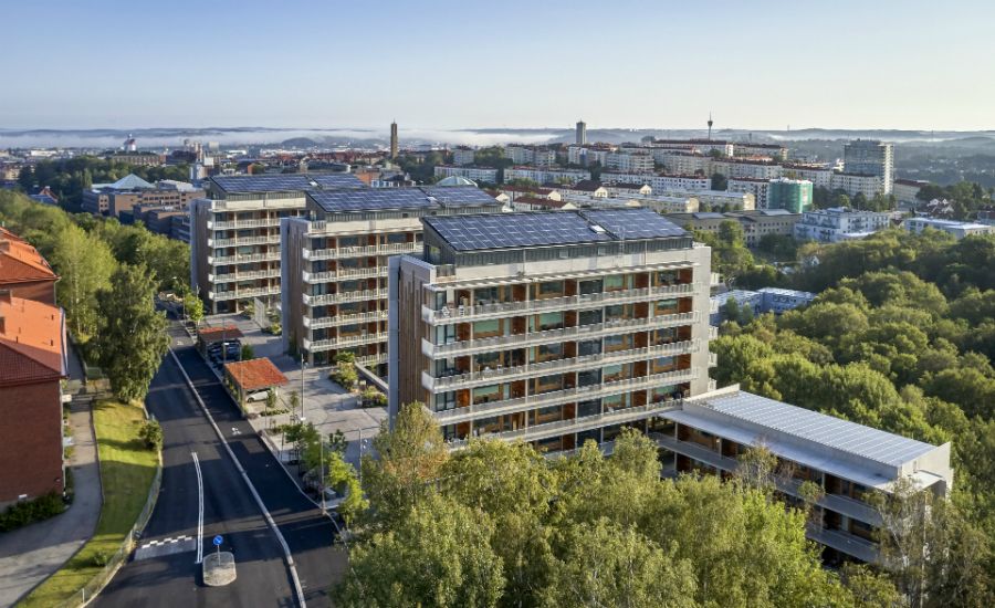 Luftfoto av moderna flerbostadshus med solpaneler på taken, omgivna av grönska med en stad i bakgrunden