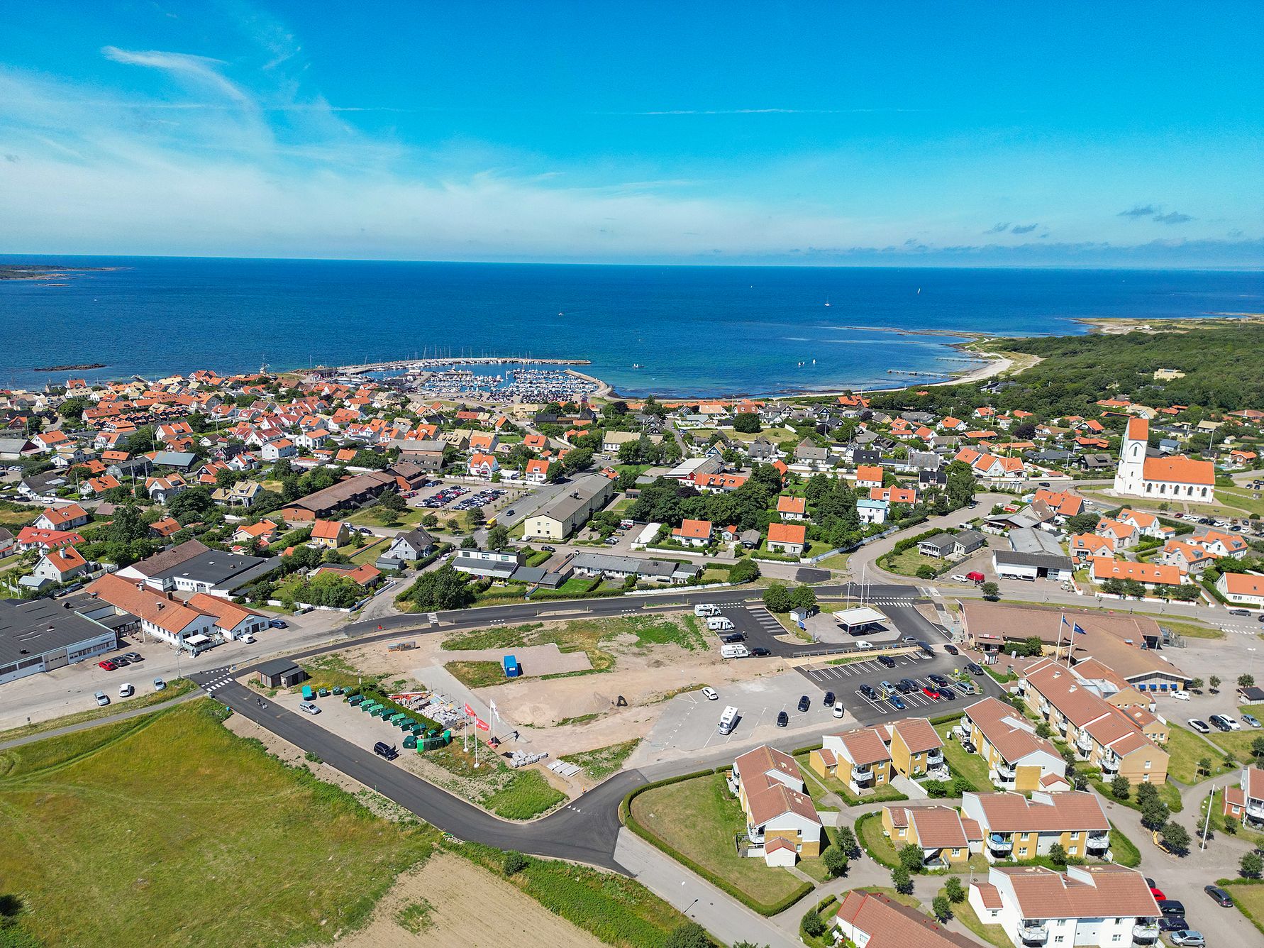 Flygfoto över en kuststad med röda tegeltak, småbåtshamn och kyrka nära havet under klarblå himmel