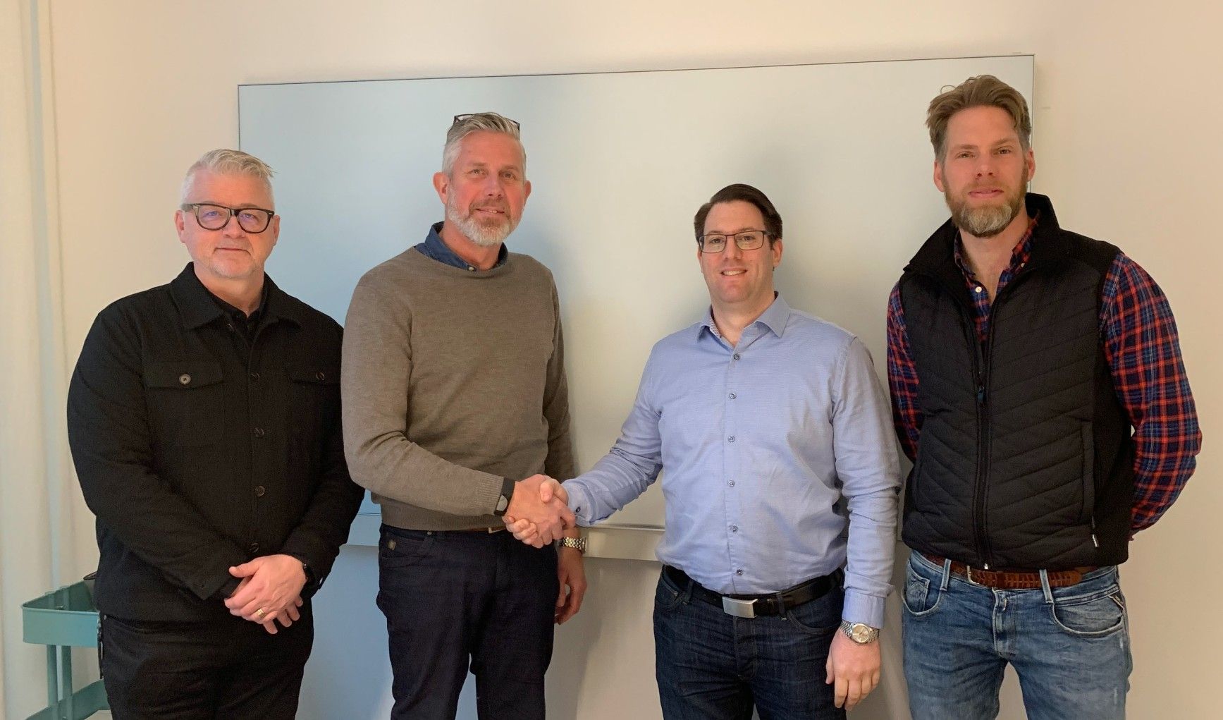 Fyra personer står framför en whiteboard, där två av dem skakar hand. De är klädda i avslappnad kontorsklädsel