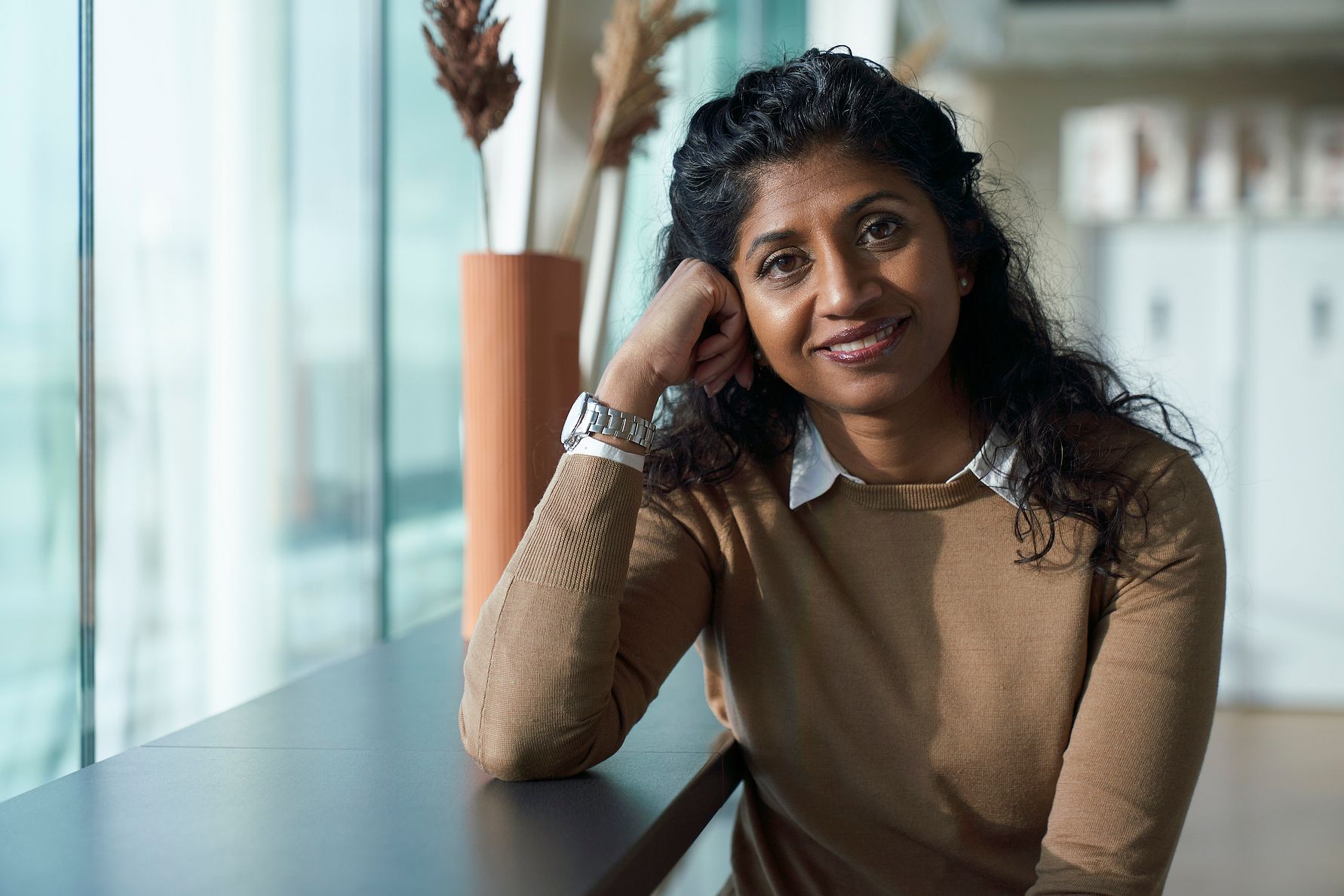 Kvinna i brun tröja med vit krage sitter vid ett bord bredvid ett stort fönster, med torkade blommor i en vas i bakgrunden