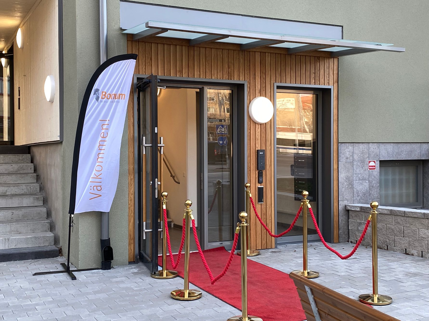 Ingång till en byggnad med röda rep, en röd matta, och en flagga med texten "Välkommen" vid sidan
