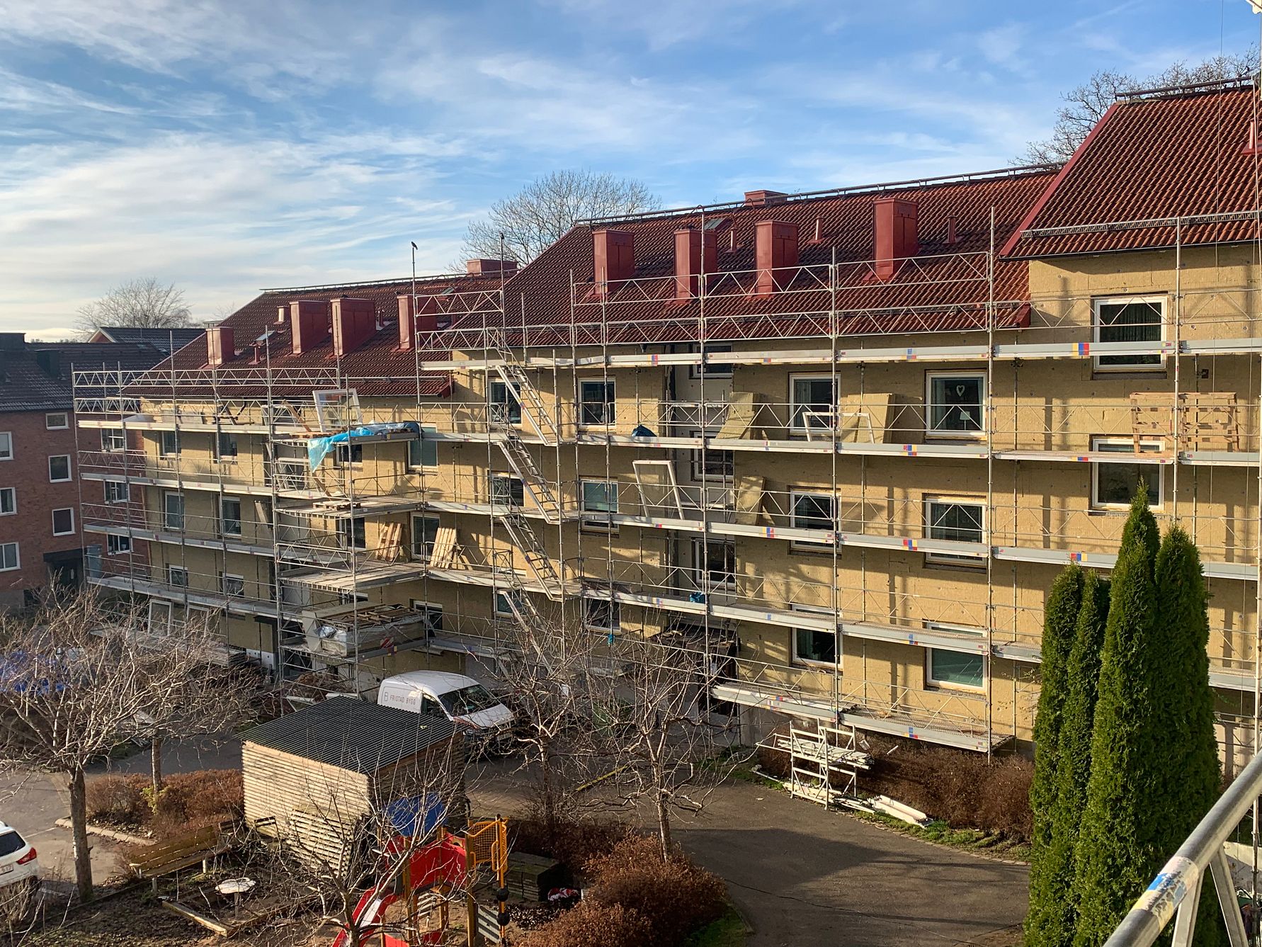 Flerbostadshus under renovering med byggställningar och skyddsnät. Soligt väder med blå himmel och träd i förgrunden