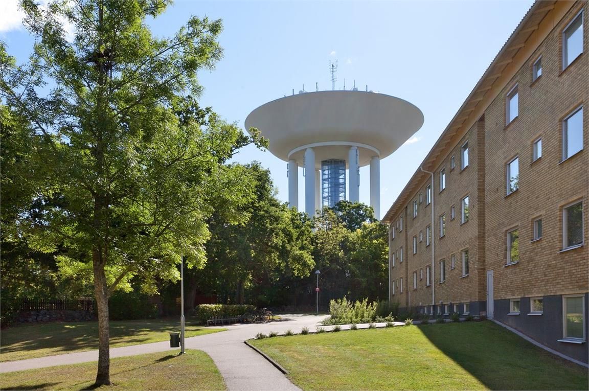 Vattentorn omgivet av träd och grönska, med ett bostadshus i tegel till höger och en gångväg i förgrunden