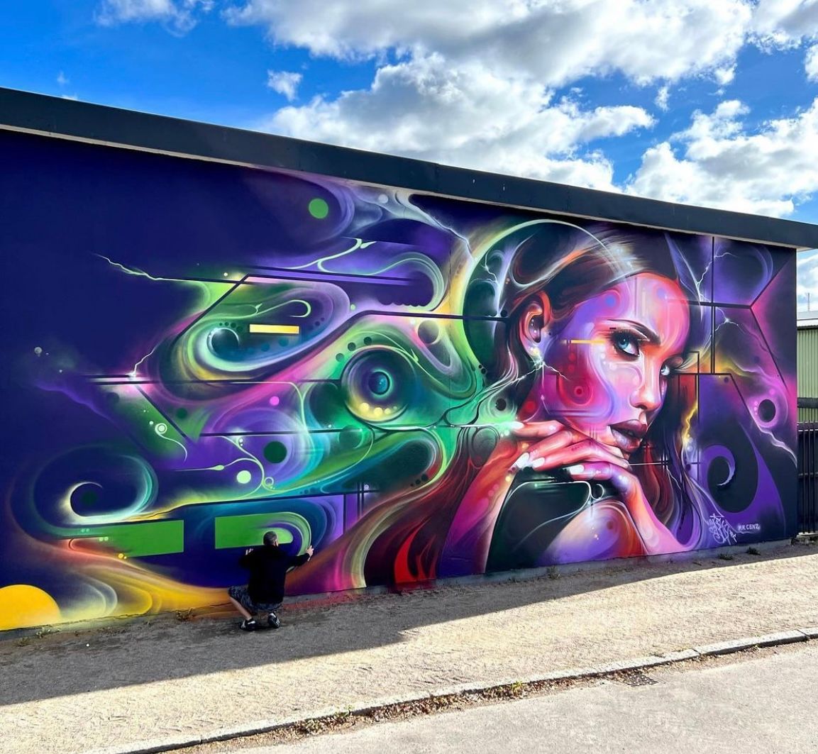 Väggmålning i färgglada abstrakta motiv med svarta, gröna, lila och gula färger på en byggnad under en blå himmel med moln. En person sitter på marken framför väggen och målar