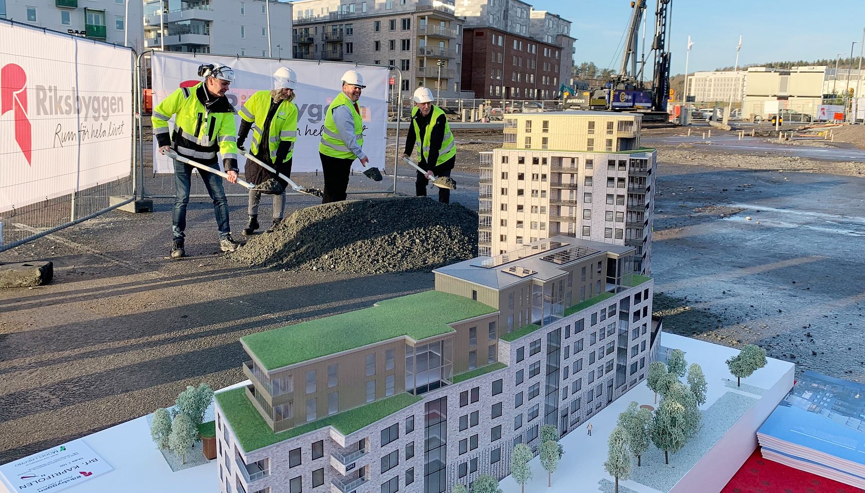 Personer i reflexkläder utför en ceremoniell första spadtag vid en byggarbetsplats, med en arkitektonisk modell av ett bostadsprojekt i förgrunden. Bakgrunden visar flerfamiljshus och byggutrustning