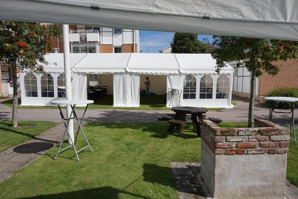Vit tältpaviljong uppställd på en grön gräsmatta, omgiven av bord, träd och byggnader i bakgrunden