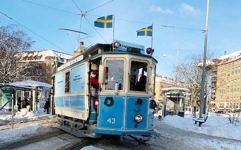 Blå spårvagn med nummer 43 kör genom ett snöigt stadsmiljö, med svenska flaggor ovanpå och passagerare ombord