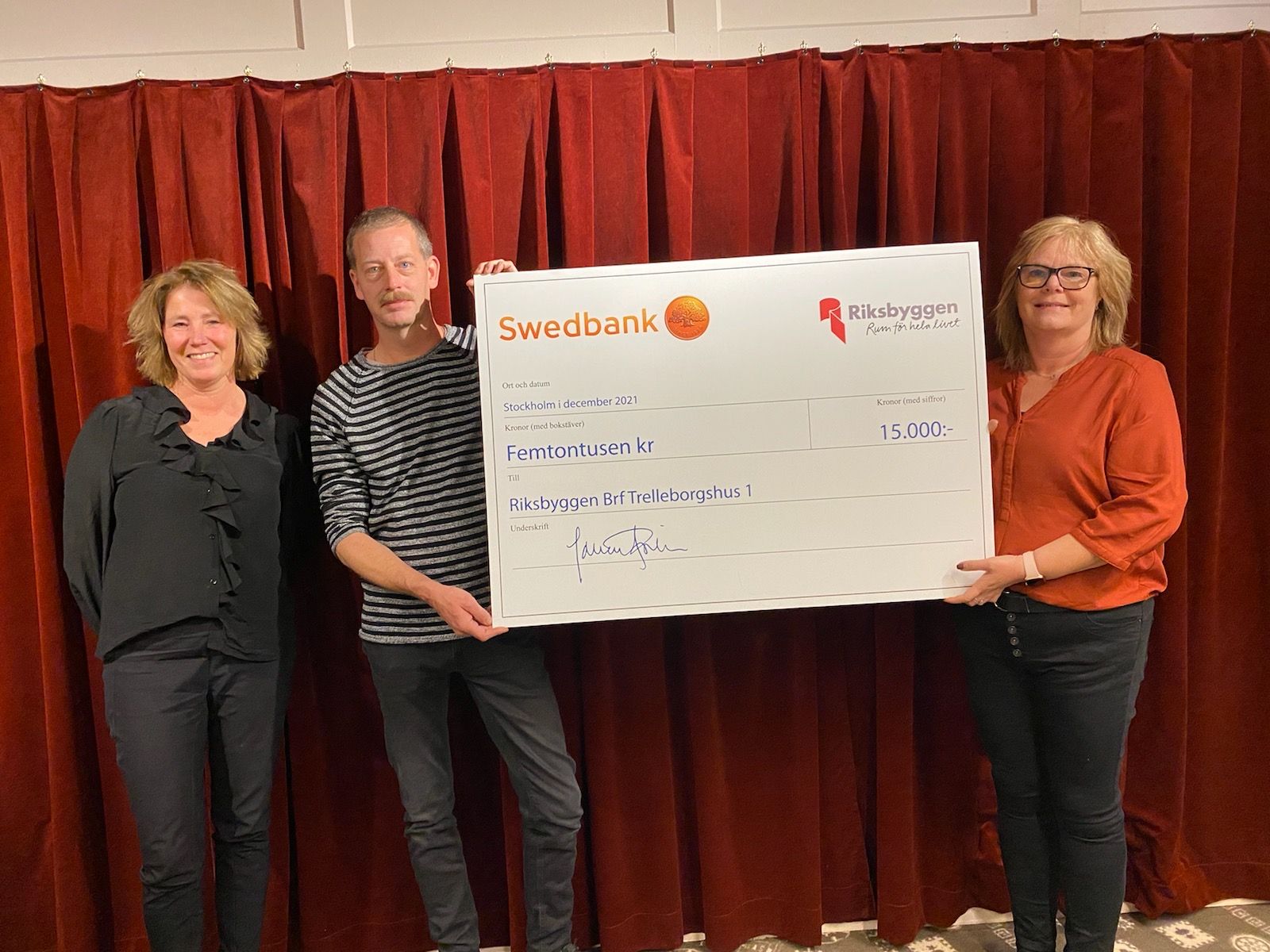 Fyra personer står framför ett rött draperi och håller upp en stor symbolisk check med texten "Femtonusen kr" från Swedbank och Riksbyggen