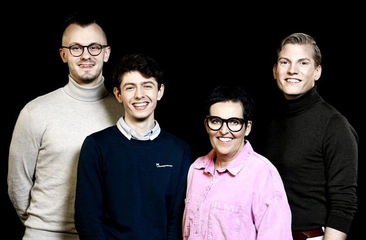 Fyra personer står framför en svart bakgrund, klädda i olika tröjor och skjortor i ljusa och mörka färger